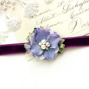 May include: A purple velvet choker with a fabric flower in shades of blue and lavender, adorned with pearls. The flower sits on a green ribbon base. The choker is set against a vintage postcard.