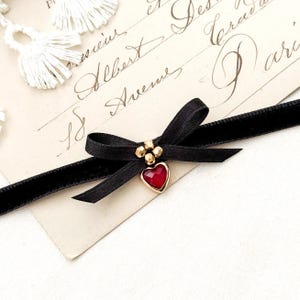 May include: A black velvet choker necklace with a bow and a heart-shaped pendant. The pendant has a red gemstone set in a gold-colored frame. The bow is centered on the choker, and the pendant hangs below it. The background includes a vintage paper with script and white tassels.