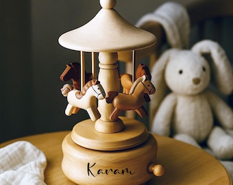 May include: A wooden carousel music box with four carved horses in shades of brown and cream. The carousel has a light-colored wooden canopy and base. The name "Karam" is engraved on the base. A pacifier and a stuffed bunny are in the background.