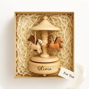 May include: A wooden carousel music box with four carved horses in a gift box. The carousel is light brown with a domed roof and a base that reads "Olivia". A tag that says "For You" is attached.