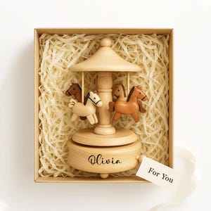 May include: A wooden carousel music box with four carved horses in a gift box. The carousel is light brown with the name "Olivia" engraved on the base. A tag that says "For You" is attached with a white ribbon.