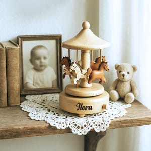 May include: A wooden carousel music box with four horse figurines in shades of brown and cream. The base is engraved with the name "Flora". A framed baby photo, a teddy bear, and books are on a wooden shelf.