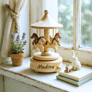 May include: A wooden carousel music box with four horses, a domed roof, and the name "Medina" engraved on the base. The carousel is light brown, and the horses have brown manes. A small white rabbit figurine and a stack of books are nearby.