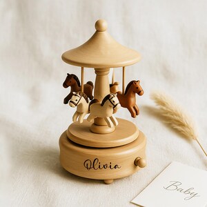 May include: Wooden carousel music box with four carved horses in shades of brown and cream. The carousel has a light brown, domed roof and a base with the name "Olivia" engraved. A small card with the word "Baby" is visible.
