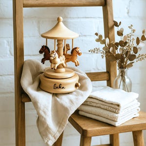 May include: A wooden carousel music box with four horse figurines in shades of brown and cream. The base of the carousel is engraved with the name "Luka". The carousel sits on a wooden chair with a beige cloth draped over it. A vase of dried flowers is in the background.