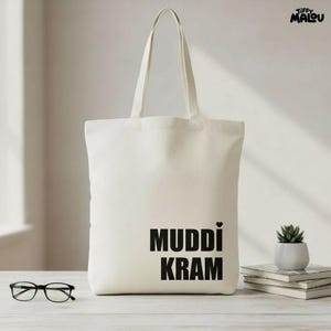 Könnte beinhalten: Cremefarbene Tragetasche mit schwarzem Text „MUDDI KRAM“ und einem Herz. Die Tasche hat lange Henkel. Eine Brille und eine kleine Topfpflanze sind ebenfalls auf einer weißen Oberfläche zu sehen.