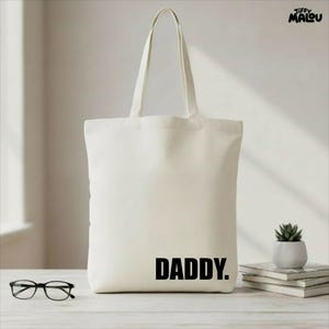 Puede incluir: Bolso tote de color crema con la palabra "DADDY" impresa en letras negras en negrita. El bolso tiene asas largas. También se ven unas gafas y una pequeña planta en maceta. El bolso está sobre una superficie clara.