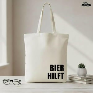 Pode incluir: Saco tote creme com a frase "BIER HILFT" em preto. O saco tem alças compridas e está sobre uma superfície clara. Também são visíveis uns óculos e uma pequena planta em vaso.