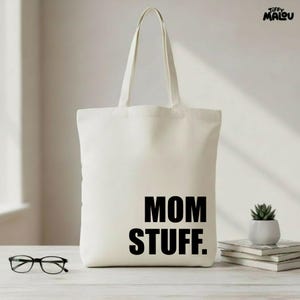 Puede incluir: Bolso tote de color crema con las palabras "MOM STUFF." impresas en letras negras en negrita. El bolso tiene dos asas y está sobre una superficie clara. También se ven unas gafas y una pequeña planta en maceta.