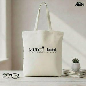 Könnte beinhalten: Cremefarbene Tragetasche mit langen Henkeln. Auf der Tasche steht in schwarzer Schrift "MUDDI x Beutel HIGH SOCIETY CLUB". Eine Brille und eine kleine Topfpflanze sind im Hintergrund zu sehen.