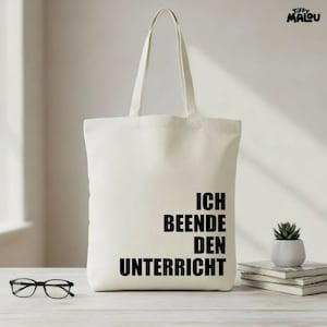 Könnte beinhalten: Cremefarbene Tragetasche mit schwarzem Text "ICH BEENDE DEN UNTERRICHT". Die Tasche hat zwei Henkel und steht auf einer hellen Oberfläche. Eine Brille und eine kleine Topfpflanze sind ebenfalls zu sehen.