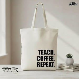 Könnte beinhalten: Beige Canvas-Tragetasche mit dem Text "TEACH. COFFEE. REPEAT." in fetten schwarzen Buchstaben. Die Tasche hat zwei Henkel und steht auf einer hellen Oberfläche. Eine Brille und eine kleine Topfpflanze sind ebenfalls zu sehen.