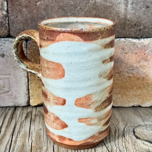 May include: Handmade ceramic mug with a unique design. The mug features a light blue and brown pattern with a textured surface. The handle is a matching brown color. This mug is perfect for coffee, tea, or any hot beverage.