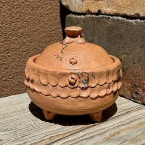 以下が含まれることがあります： 蓋付きのテラコッタ製の壺で、縁が波打っていて、小さな足が3本ついています。壺は茶色の釉薬がかかっており、黒い模様があります。