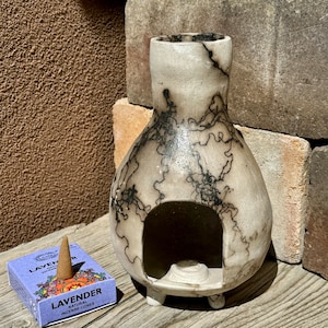 以下が含まれることがあります： ユニークな丸みを帯びた形状と3つの小さな足を持つセラミック香炉。オフホワイトの表面には、印象的な黒い稲妻模様があります。「LAVENDER」の文字が見えるラベンダーの香りのコーンの箱が香炉の隣にあります。