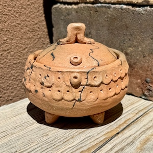 以下が含まれることがあります： 蓋付きの小さなテラコッタ色の陶器の壺。壺は黒の線と小さな穴のあるテクスチャのある表面をしています。蓋は2本の脚を持つユニークな3次元デザインです。