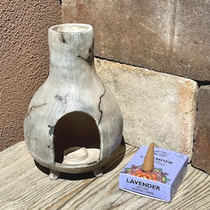 以下が含まれることがあります： 大きな開口部と3つの小さな足が付いた灰色のセラミック香炉。ラベンダーのお香コーンの箱と単一のコーンがバーナーの隣にあります。バーナーには濃い灰色のひび割れ模様があります。