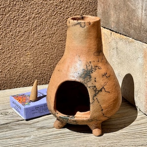 以下が含まれることがあります： 大きな開口部と3つの小さな足が付いた、茶色のセラミックチムニー。チムニーには黒いマーキングがあり、ラベンダーのお香の箱と円錐形のお香の隣にあります。背景はテクスチャード加工された壁と木製の表面です。