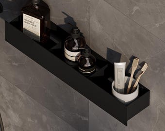 May include: A matte black rectangular shower shelf mounted on a gray tiled wall. The shelf holds two dark brown pump bottles, a small dark bottle, and a white ceramic cup containing two wooden toothbrushes and a tube of toothpaste.