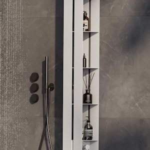 May include: A tall, vertical white metal shower shelf unit featuring multiple open compartments for storage. The unit is mounted on a wall and holds various bottles, a reed diffuser, and a round beige scrub brush hanging from a bottom hook.