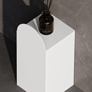 Puede incluir: Un pedestal blanco con un borde superior en arco redondeado. Sobre la superficie plana hay un frasco difusor de vidrio marrón oscuro con varillas negras. El frasco tiene el texto ETERNAL MUSK impreso en blanco en el vidrio.