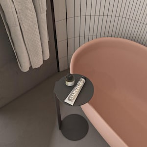 May include: A dark gray metal side table with a circular top and base, holding a small dark bottle and a tube of Marvis toothpaste. The table is positioned next to a smooth, light pink bathtub and a light gray textured bathrobe hanging nearby.