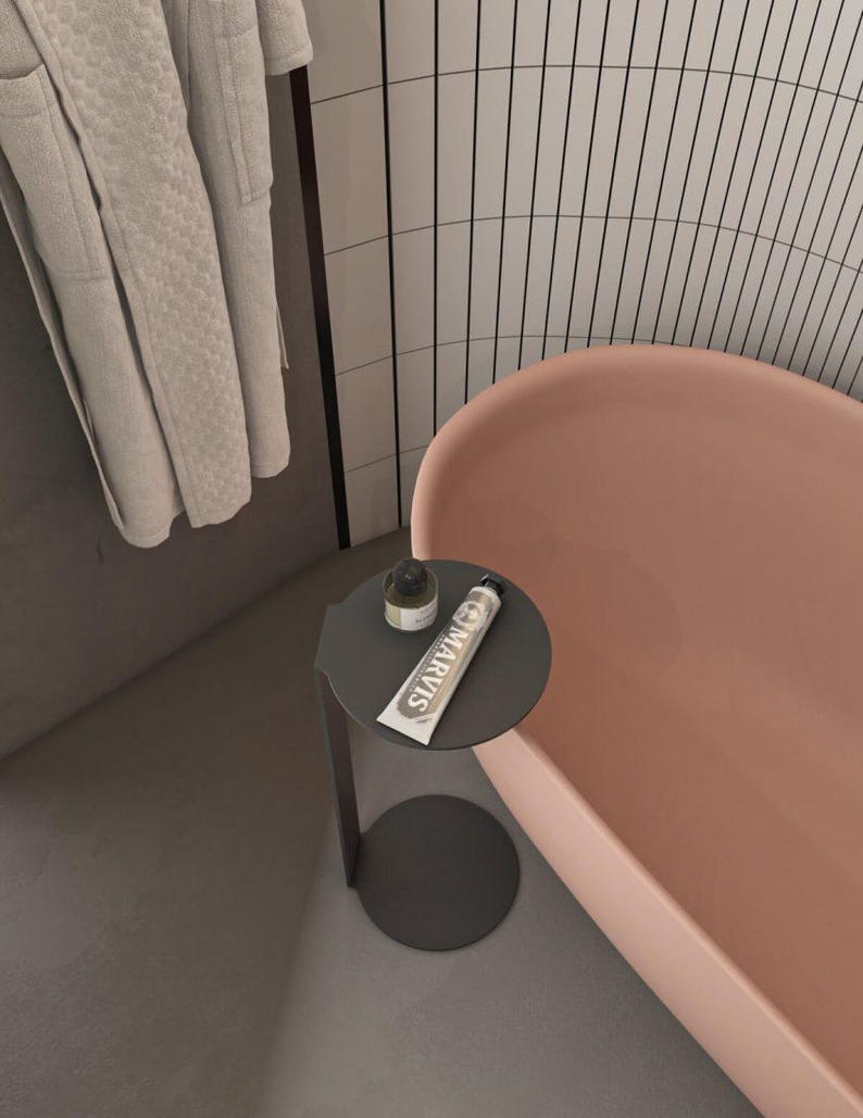 May include: A dark gray metal side table with a circular top and base, holding a small dark bottle and a tube of Marvis toothpaste. The table is positioned next to a smooth, light pink bathtub and a light gray textured bathrobe hanging nearby.