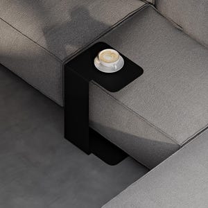 May include: A matte black C-shaped side table with a smooth, flat surface positioned over a grey sofa. A white ceramic cup and saucer containing a latte with heart-shaped foam art sits on the table, which has a thin, vertical metal base.