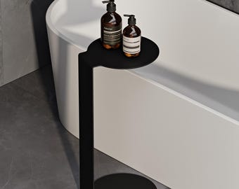 May include: A matte black metal side table with a circular top and base, connected by a single vertical support. Two amber-colored glass pump bottles with black dispensers and white labels reading Aesop are placed on the circular tabletop surface.