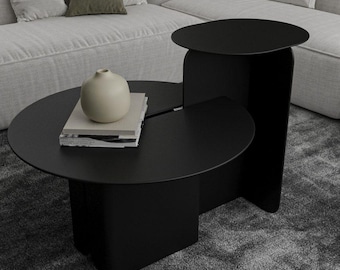 May include: A modern black coffee table with a unique design. The table features two circular tabletops connected by a curved support, and a smaller side table. A beige vase and books are on the table, set against a gray carpet and a white sofa.