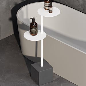 May include: A white metal tiered stand with two circular shelves supported by a central pole and a heavy dark gray cubic base. The stand holds three amber-colored glass bottles with black pump dispensers and labels reading Aesop.
