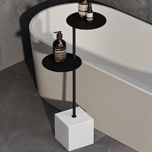 May include: A black metal stand with two circular shelves and a white cubic base. The stand holds three amber-colored glass bottles with black pump dispensers, featuring labels with the text Aesop. The structure is vertical and tiered.