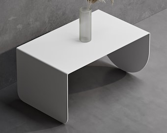 May include: A white coffee table with a smooth, continuous curved design that forms the legs and top. A clear, vertically ribbed glass vase sits on the surface, containing three dried stalks of pampas grass and wheat-like decorative stems.