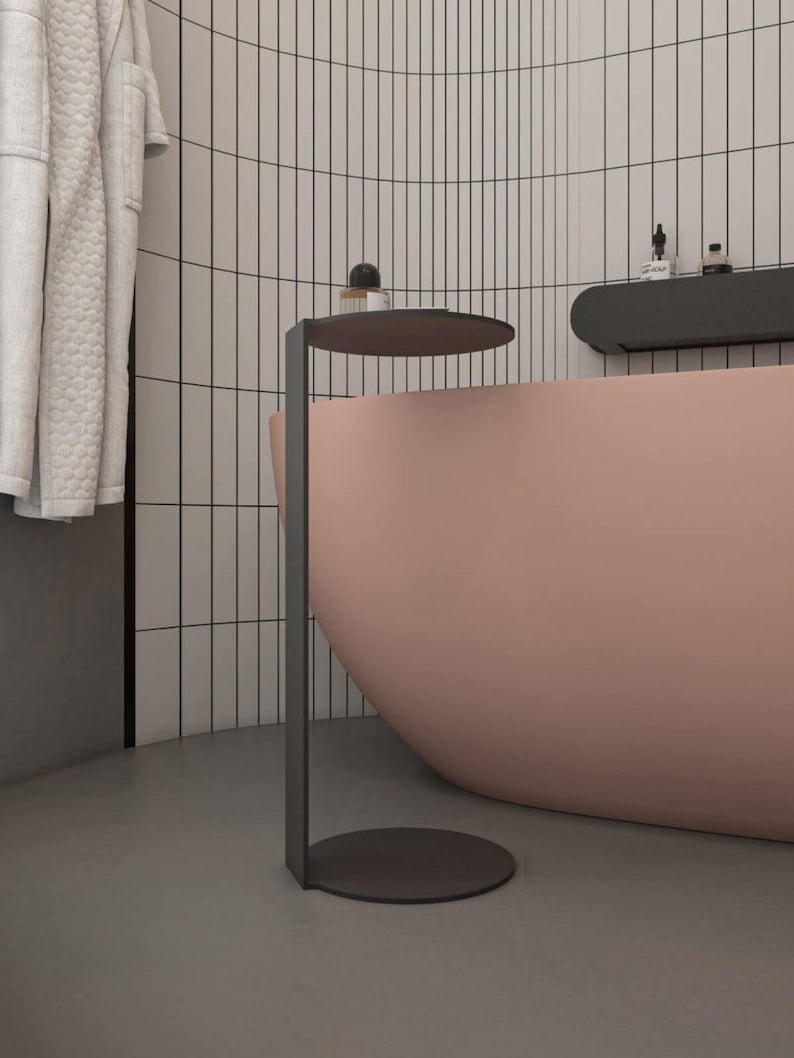 May include: A matte black metal side table featuring a slender vertical support, a circular base, and a matching circular top surface. The table is positioned next to a curved, solid light pink bathtub in a bathroom with white tiled walls.