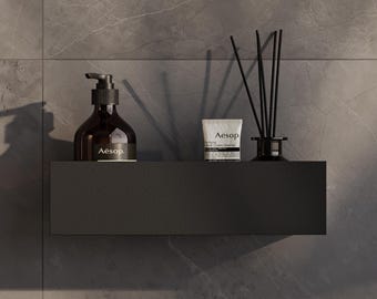 May include: A matte black rectangular wall-mounted shelf holding an amber glass bottle with a black pump dispenser labeled Aesop, a white tube of Aesop Purifying Facial Cream Cleanser, and a black glass reed diffuser with black reeds.
