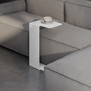 May include: A white C-shaped side table made of a single continuous piece of metal. The table features a flat rectangular top surface and a vertical base that slides under furniture. A white ceramic cup and saucer are placed on the table top.