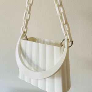 Puede incluir: Un bolso blanco con una correa de cadena y un asa curva. El bolso tiene un diseño vertical acanalado y está suspendido de una cadena blanca. El bolso está hecho de un material sólido y opaco.