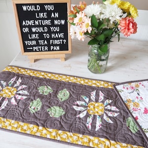 Może przedstawiać: Szary pikowany bieżnik stołowy z żółtą obwódką i kwiatowym wzorem. Bieżnik zawiera cytat z Piotrusia Pana: "Chcesz teraz przeżyć przygodę, czy wolisz najpierw napić się herbaty?"