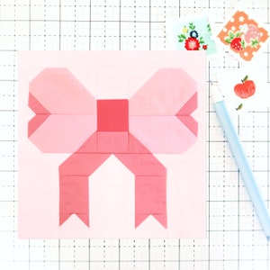 Peut inclure: Un nœud en tissu rose avec un centre rouge, cousu sur un fond rose. Le nœud est composé de plusieurs carrés de tissu cousus ensemble.