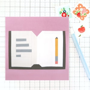 May include: A pink background with a white notebook and a yellow pencil. The notebook has gray lines and a black spine. The pencil has a gray eraser and a red tip.