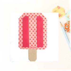 May include: A pink and white popsicle with a brown stick. The popsicle is made of fabric and has a polka dot pattern.