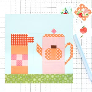 May include: A patchwork quilt block featuring a pink teapot with orange floral accents and a matching stack of cups. The design includes a green floral border at the bottom and a light blue background. A light blue pen is in the image.