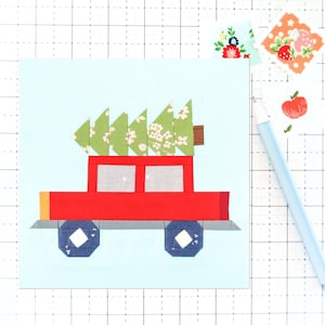 Pode incluir: Um caminhão de brinquedo vermelho com uma árvore de Natal verde no topo. O caminhão tem faróis amarelos, janelas cinzas e rodas azuis. A árvore é feita de tecido verde com flores brancas.