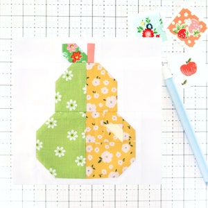 May include: A patchwork pear quilt block made from green and yellow fabric with a white floral pattern. The block is made up of several smaller squares and triangles sewn together.