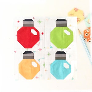 May include: A square quilt block featuring four colorful Christmas light bulb designs. Each bulb is a different color: red, green, orange, and blue. The bulbs have gray tops and white highlights, set against a white background with star patterns.