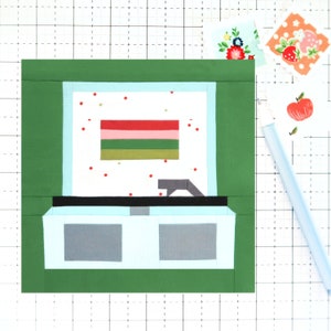 May include: A green framed quilt block with a white background and a red, pink, green, and gold striped rectangle in the centre. The background is covered in small red dots. The bottom of the block has a black rectangle with a grey rectangle on top of it. The block is surrounded by a white grid.
