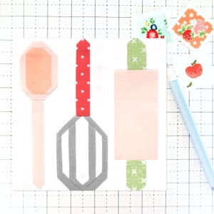 May include: Three fabric kitchen tools, a pink spatula, a gray whisk, and a pink rolling pin, all with fabric handles. The tools are on a white grid background.