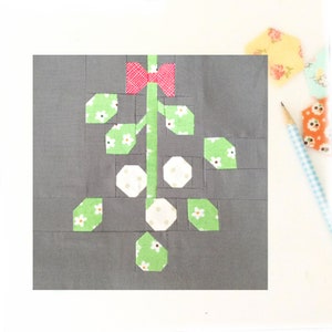 May include: A fabric patchwork block featuring a stylized floral design. The stem is green with a red bow at the top. The leaves are green with white floral patterns, and the flowers are white with polka dots. The background is gray.