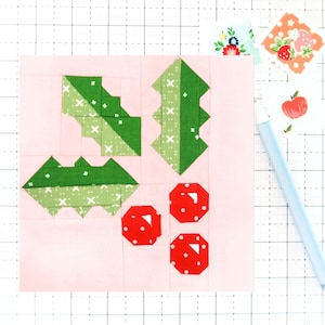 May include: A pink fabric square with a green holly leaf design and red berries. The holly leaves are made of green fabric with a white and green pattern. The berries are made of red fabric with white dots.