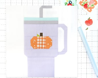 Op de afbeelding: Een paarse mok met een wit deksel en een rietje. Op de voorkant van de mok staat een kleine oranje pompoen met een wit en oranje geruit patroon.